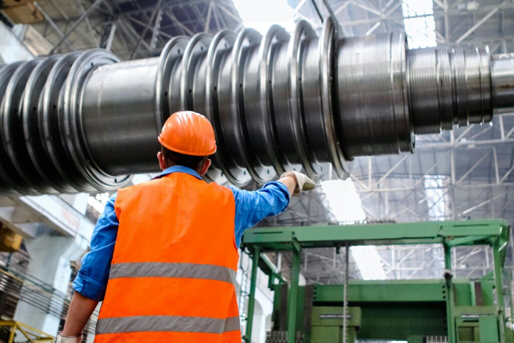 **Alt Text:** Worker in an orange safety vest and hard hat inspects large industrial machinery in a cannabis equipment manufacturing facility.